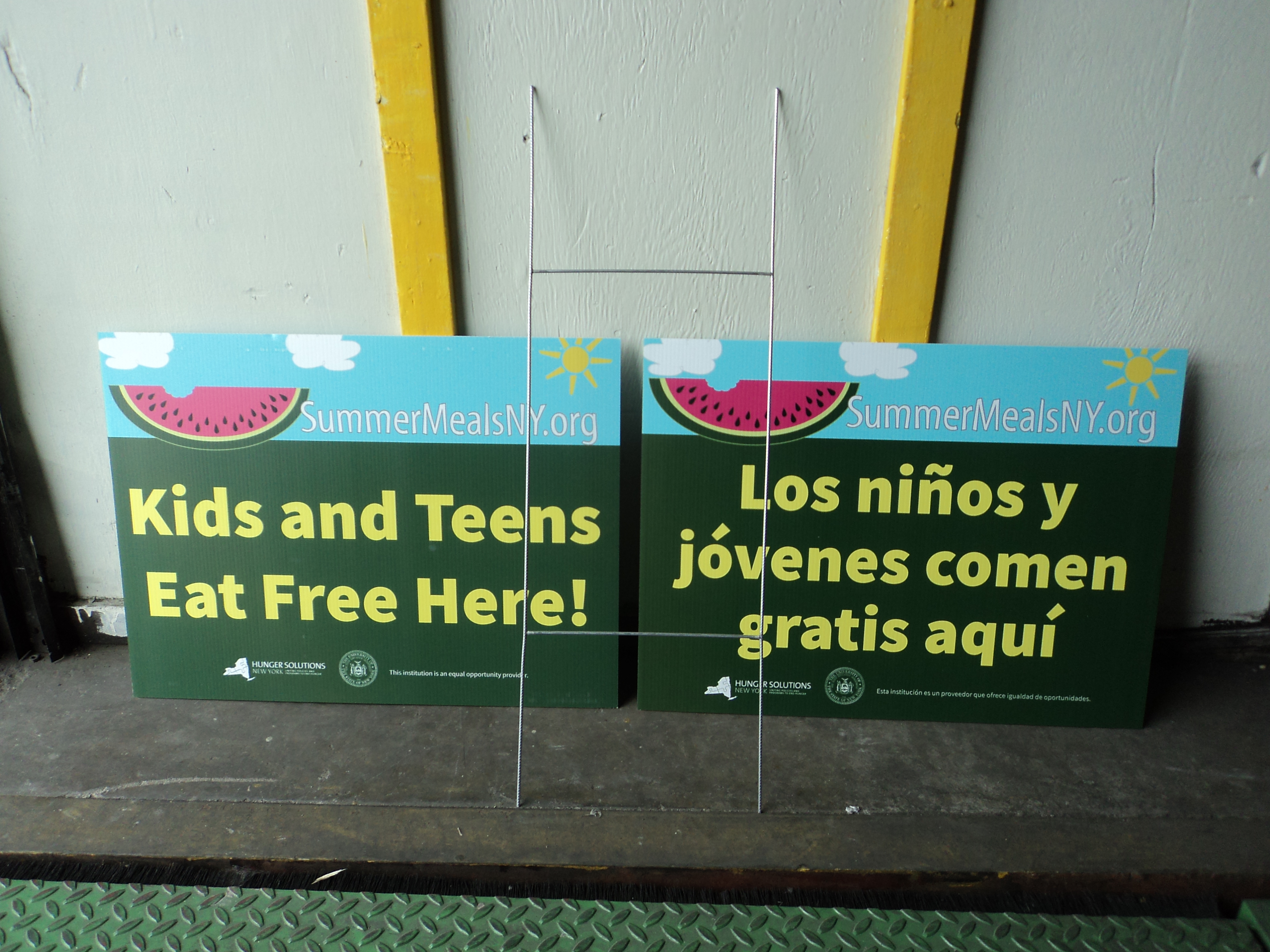 Spanish and English on one sign. SummerMealsNY.org; Kids and Teens Eat Free Here!; Hunger Solutions and NYSED logos; Spanish version on back: Los ninos y jovenes comen gratis aqui